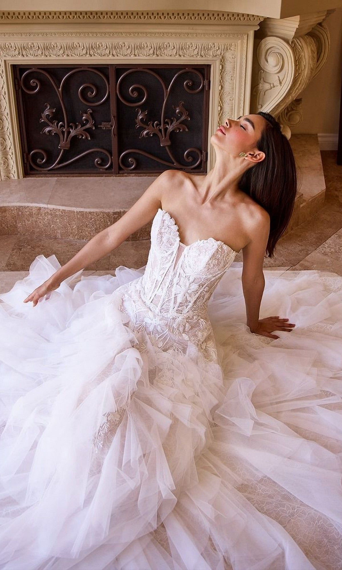 A woman wearing the Long Wedding Dress: Andrea and Leo WL016, featuring a strapless bodice and ruffle tulle a-line skirt, sits on the floor before an ornate fireplace with her head tilted upward and eyes closed.